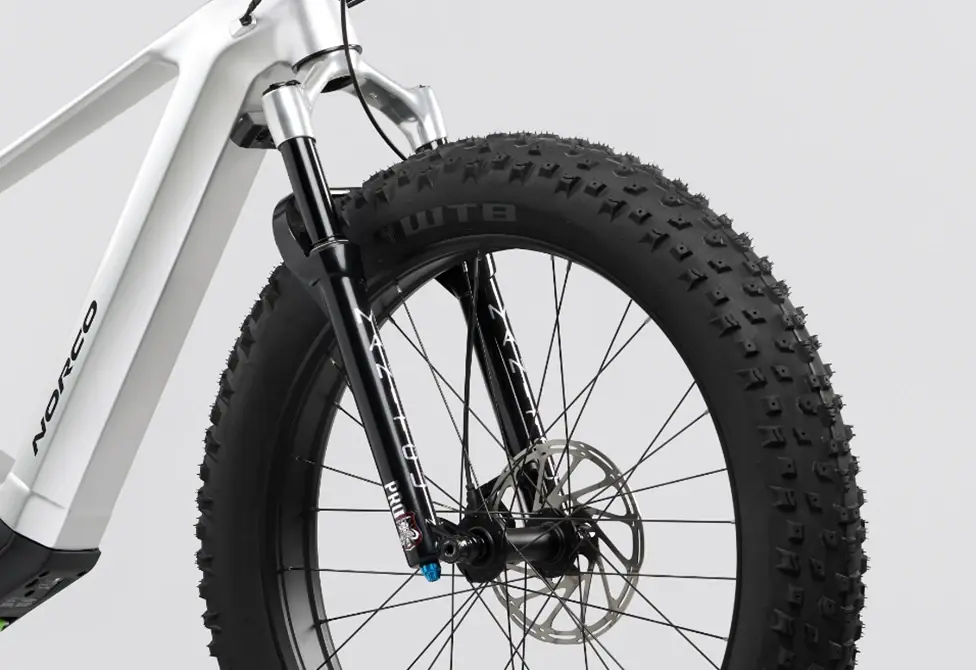 Gros plan de la fourche à suspension avant et de la roue d'un vélo blanc. Le vélo est équipé de pneus à crampons épais, d'un frein à disque et de rayons noirs. L'arrière-plan est gris clair uni.