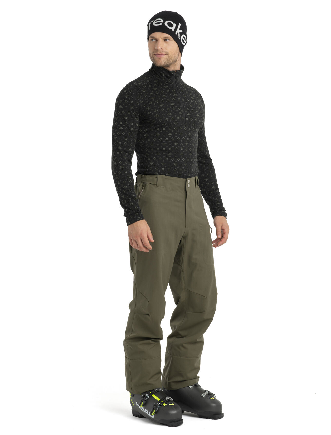 HAUT COUCHE DE BASE ICEBREAKER À MANCHE LONGUE ET DEMIS GLISSIÈRE OASIS LONG SLEEVE HALF ZIP EN MÉRINOS POUR HOMME de couleur Black/Loden vu du chandail manche longue noir avec motif hivernal des flocons de neige porter par un homme vu de face de la tête au pied avec pantalon de neige et botte de ski