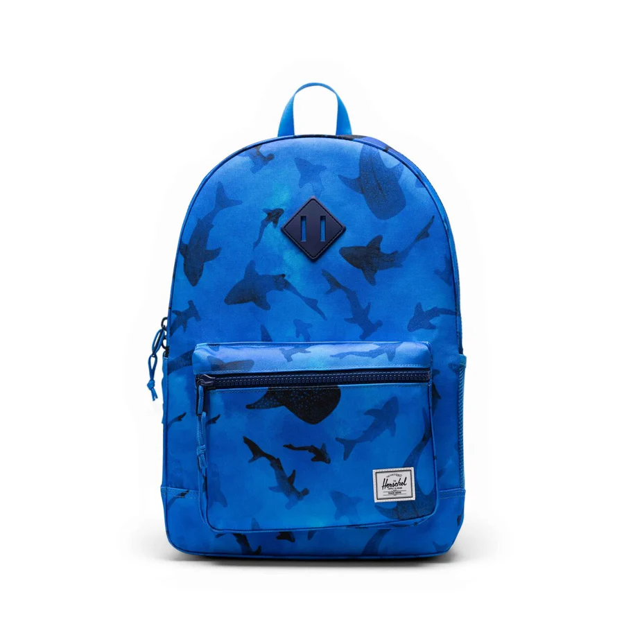Un sac à dos bleu orné d'un motif de requin, doté d'une poche avant à fermeture éclair et d'une petite étiquette blanche dans le coin inférieur droit, sur un fond blanc uni.