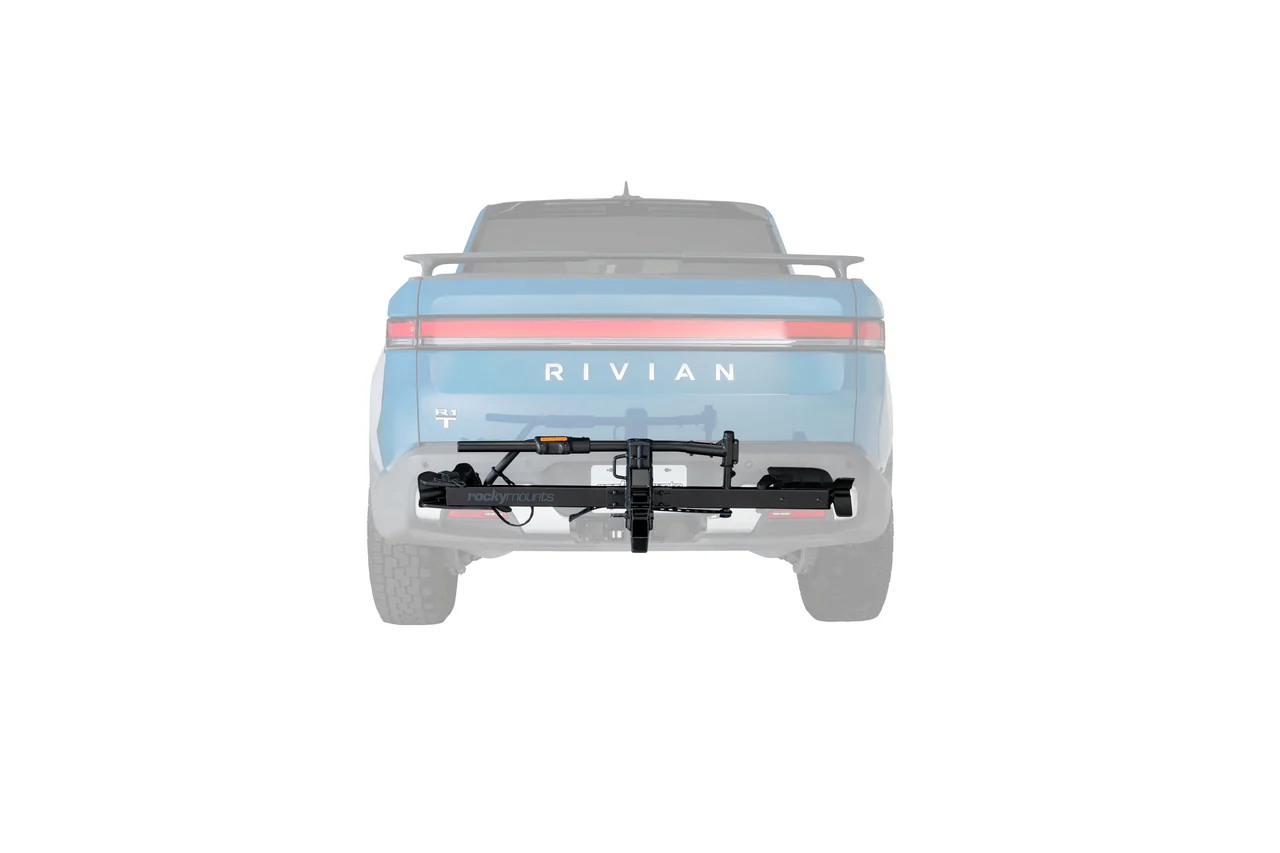 Un camion Rivian bleu est représenté à l'arrière avec un porte-vélos noir PORTE-VÉLO ROCKYMOUNTS HIGHNOON 2" (1 VÉLO) sur l'attelage de la remorque, transportant une bicyclette, le tout sur fond blanc.