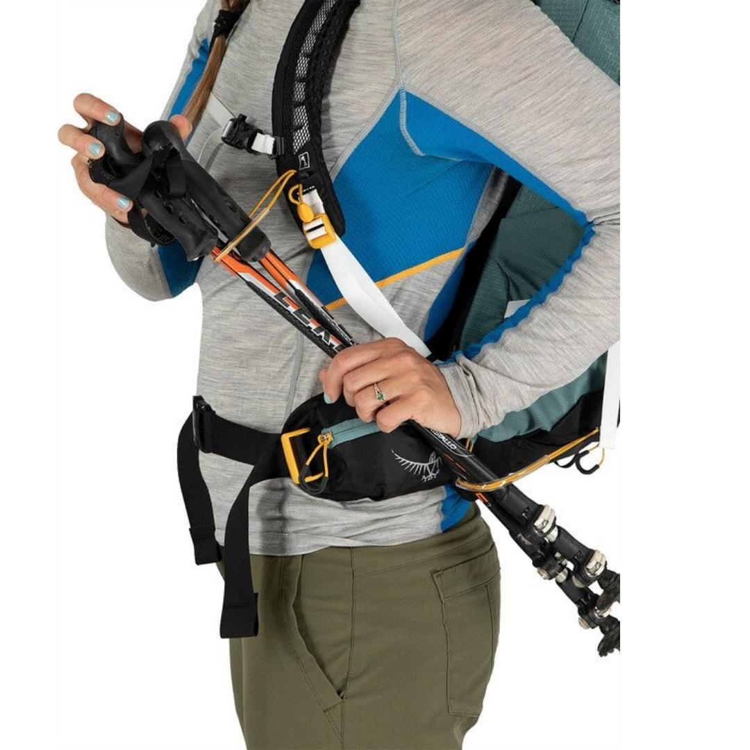 SAC À DOS OSPREY SIRRUS 24 POUR FEMME PORTÉ PAR UNE FEMME VUE DE PROFIL QUI INSTALLE DES BÂTONS DE TREKKING COURT SUR LES ATTACHE STOW-ON-THE-GO