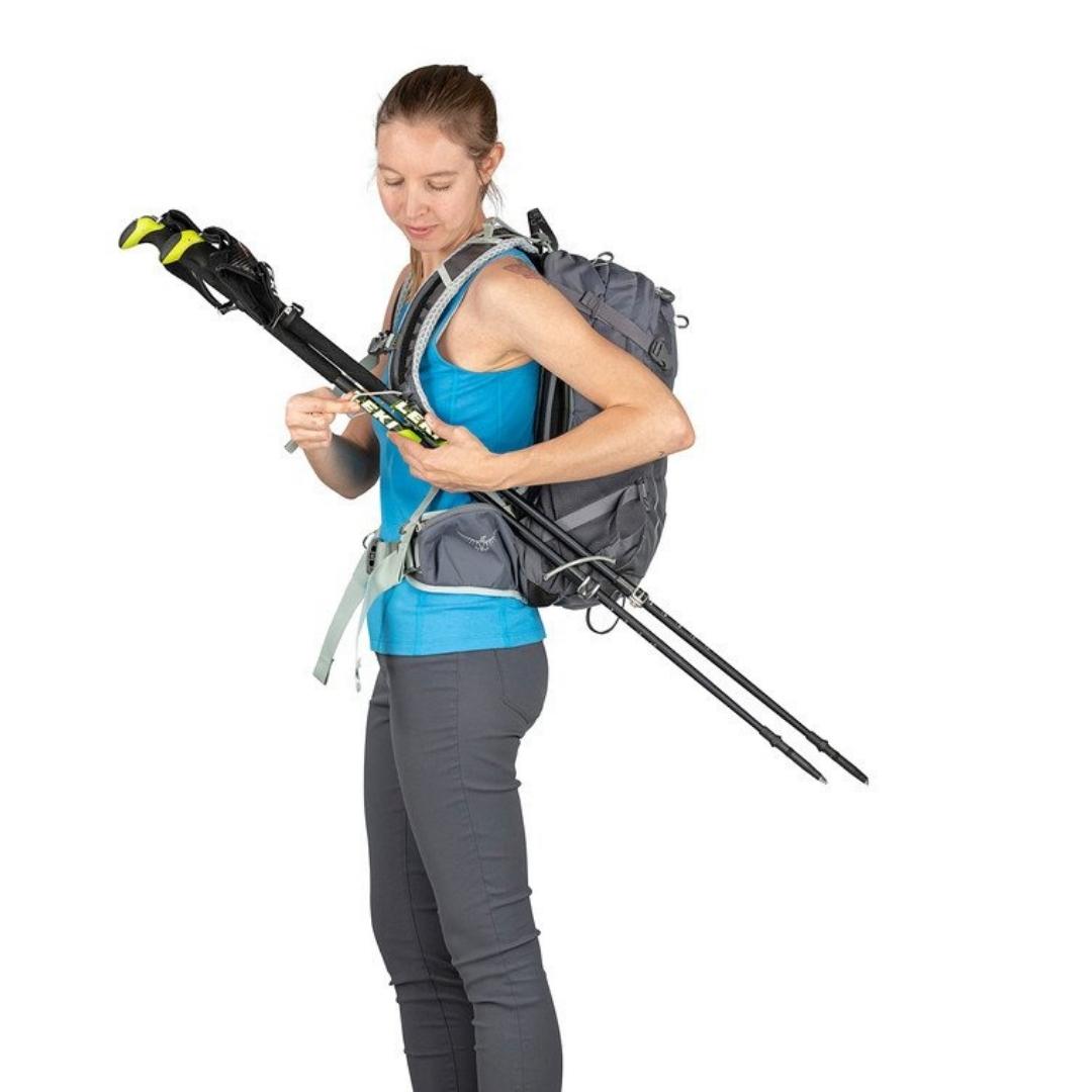 SAC À DOS OSPREY SIRRUS 24 POUR FEMME PORTÉ PAR UNE FEMME VUE DE PROFIL QUI INSTALLE DES BÂTONS DE TREKKING LONGS SUR LES ATTACHE STOW-ON-THE-GO