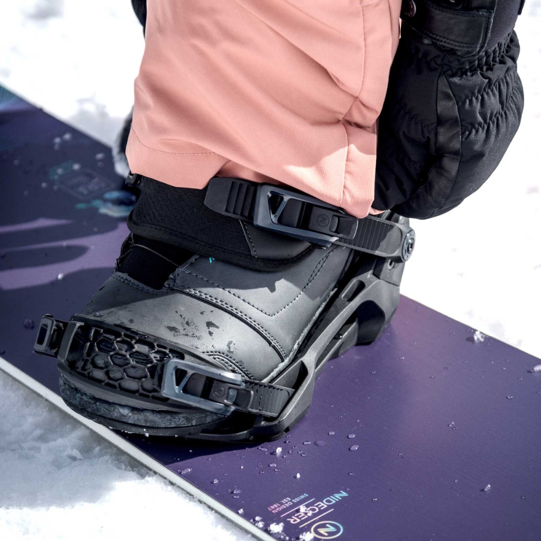 FIXATION DE PLANCHE À NEIGE NIDECKER MUON-W POUR FEMME couleur black installée sur une planche, une femme attache sa botte à la fixation