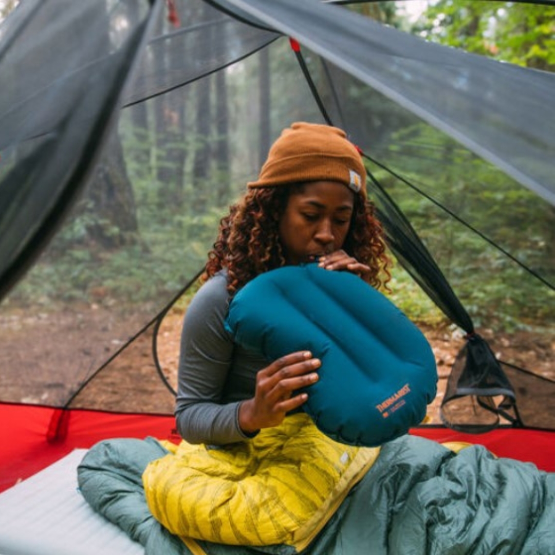 OREILLER DE CAMPING THERM-A-REST AIRHEAD LITE COULEUR DEEP PACIFIC UNE FEMME GONFLE L'OREILLER DANS SA TEMTE EN SORTIE PLEIN AIR