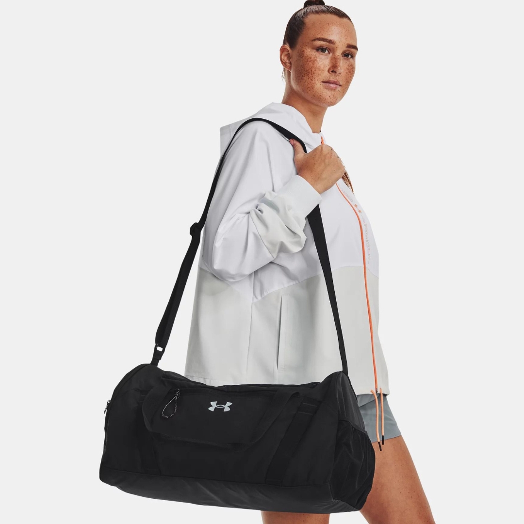 SAC DE SPORT UNDER ARMOUR UNDENIABLE SIGNATURE POUR FEMME couleur 001-BLACK/METALLIC HARBOR BLUE porté par une femme à lépaule avec la bandoulière logo under armour au haut central du sac visible