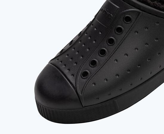 Gros plan d'une chaussure à enfiler noire avec un design perforé et sans lacets. La chaussure est dotée d'une semelle en caoutchouc et d'un embout texturé, ce qui lui donne un aspect élégant et moderne sur un fond clair.