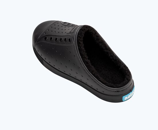 Chaussure noire de type sabot avec intérieur en peluche et fourrure. La chaussure présente un motif perforé sur la tige et une étiquette logo bleue sur le côté de la semelle.