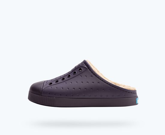 Vue de côté d'une chaussure à enfiler bleu foncé avec une semelle et un embout en caoutchouc. La chaussure présente de petites perforations et une doublure en peluche de couleur crème. Le fond est d'une couleur claire et unie.