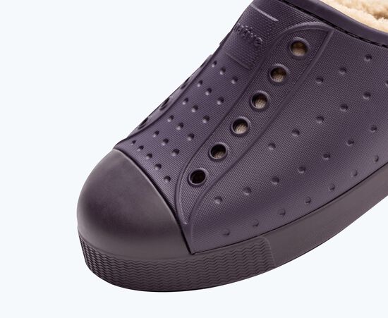 Gros plan d'une chaussure violet foncé à texture perforée, dotée de plusieurs œillets et d'un embout en caoutchouc. L'intérieur est doublé d'un matériau doux, suggérant confort et chaleur.