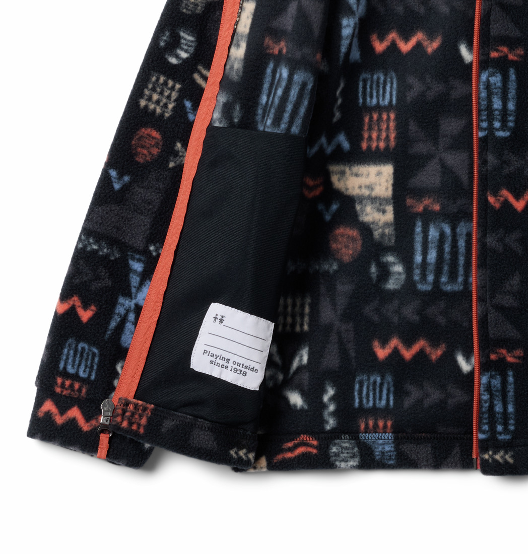 Gros plan d'une veste polaire noire zippée aux motifs géométriques colorés. L'intérieur présente une étiquette avec du texte et une petite étiquette blanche attachée. La fermeture éclair et certains éléments du motif sont de couleur orange contrastante.