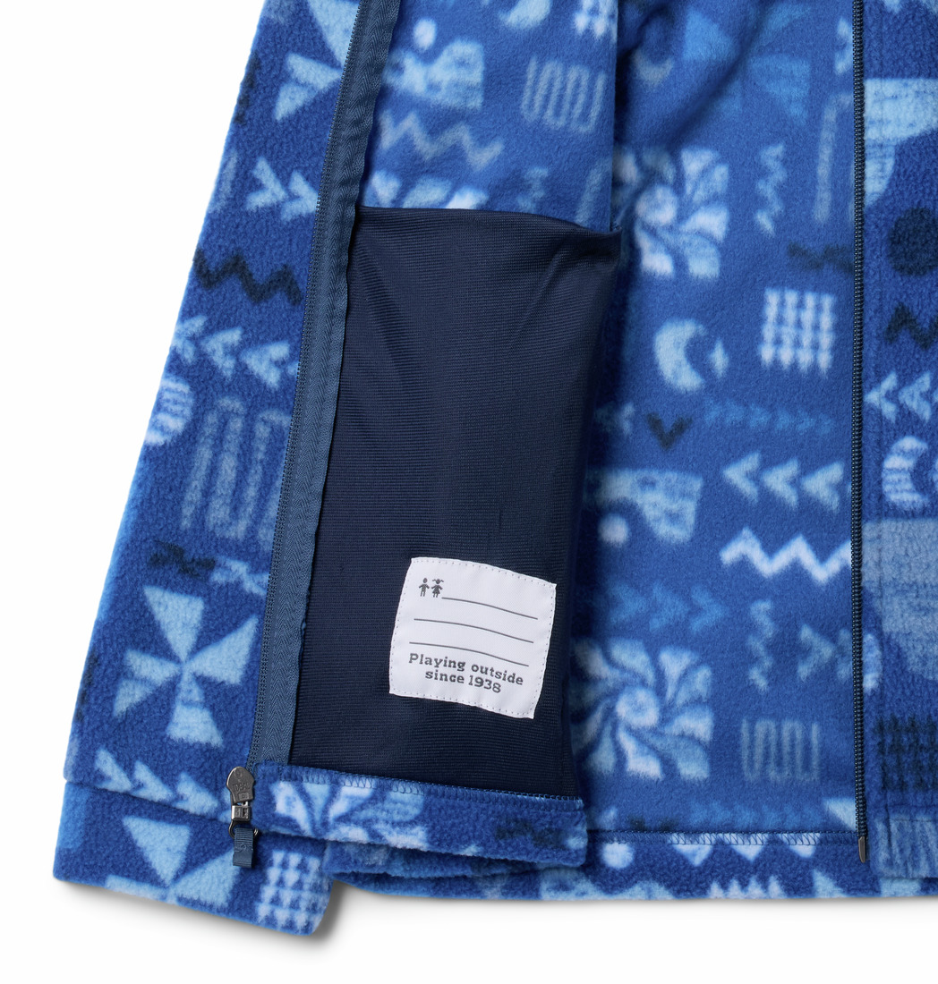 Gros plan d'une veste polaire bleue à motifs géométriques et naturels. La veste est ouverte, révélant une poche intérieure sombre et une étiquette blanche avec le texte « Jouer dehors depuis 1938 ».