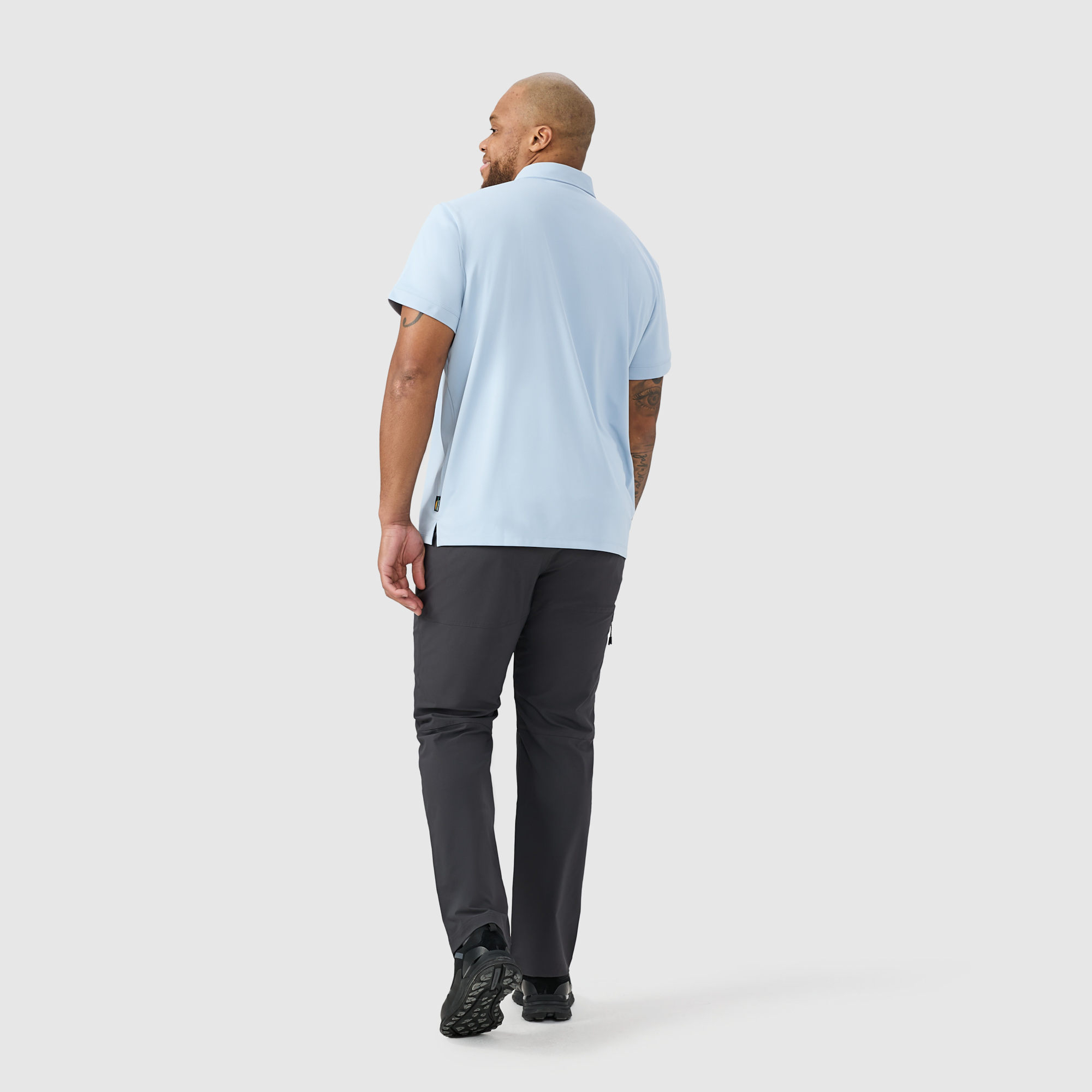 Un homme portant un polo bleu clair et un pantalon foncé s'éloigne de la caméra sur un fond gris clair uni.