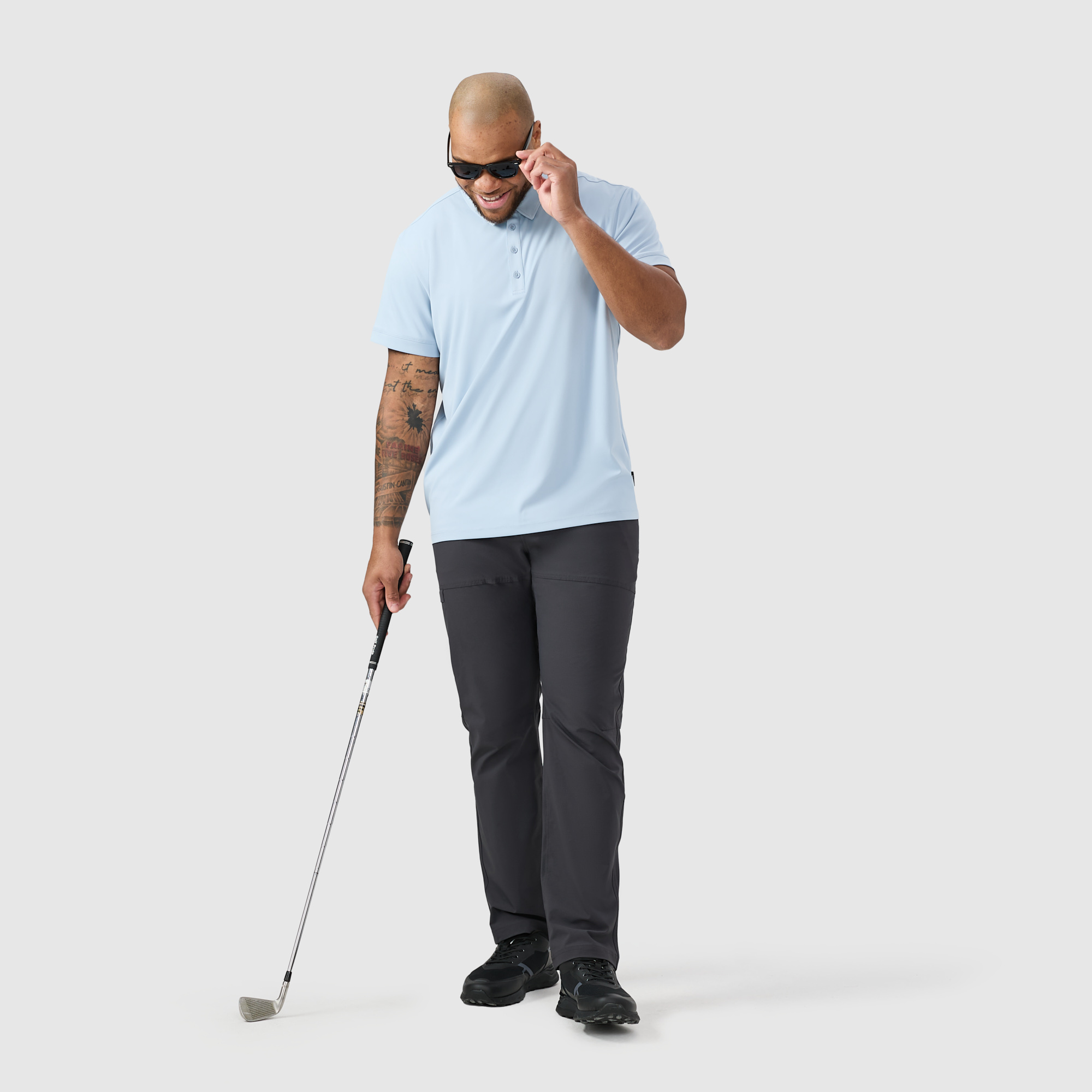 Un homme portant des lunettes de soleil, un polo bleu clair et un pantalon foncé se tient debout, tenant un club de golf, souriant et regardant vers le bas, sur un fond clair uni.