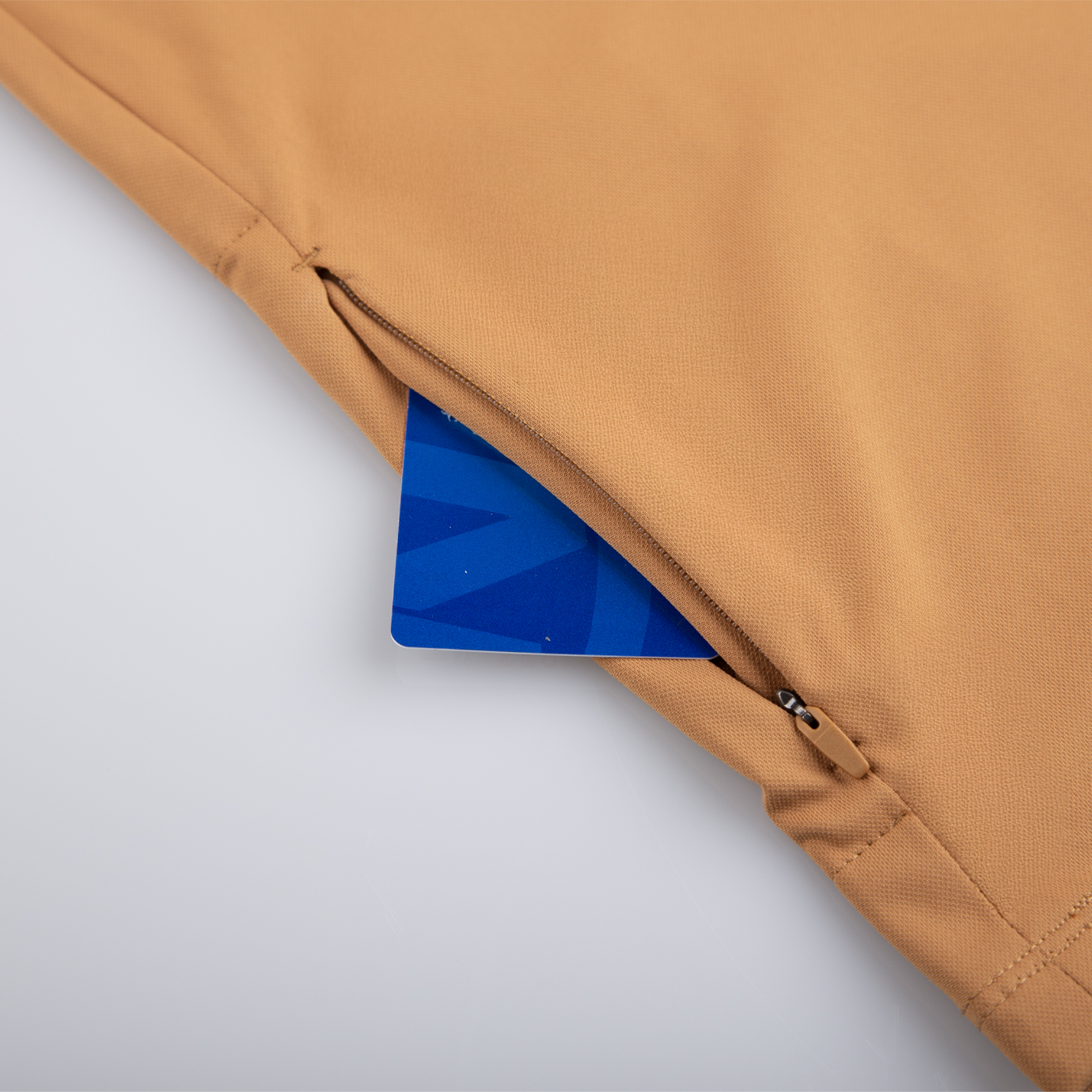 Gros plan d'un vêtement beige avec une poche zippée, partiellement ouverte pour révéler une carte bleue à l'intérieur. Le tissu paraît lisse et la fermeture éclair est petite et métallique.