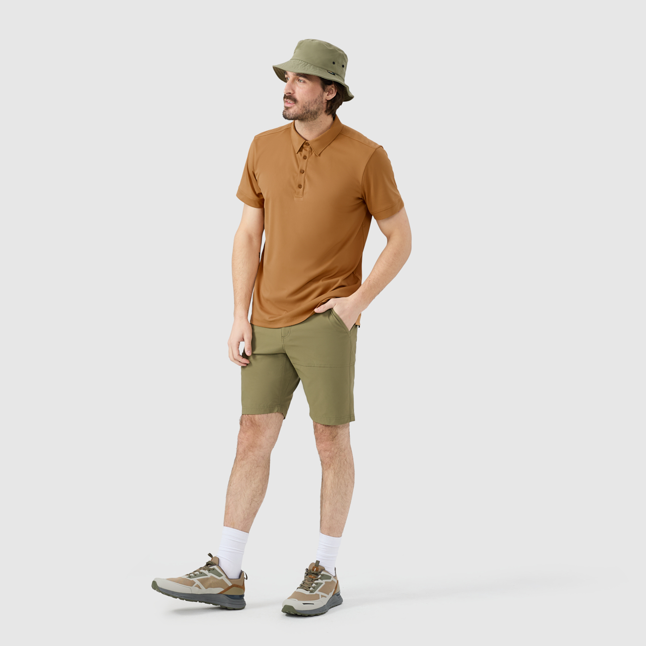 Homme debout sur un fond uni, vêtu d'un polo marron, d'un short vert olive, d'un bob vert, de chaussettes blanches et de baskets. Il a une main dans sa poche et regarde de côté, l'air détendu.