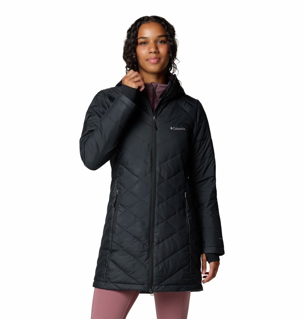 Une femme portant une longue veste Columbia matelassée noire avec une fermeture éclair et se tenant debout avec une main près du col. Elle a les cheveux bouclés et est vêtue d'un legging rose sur un fond blanc uni.