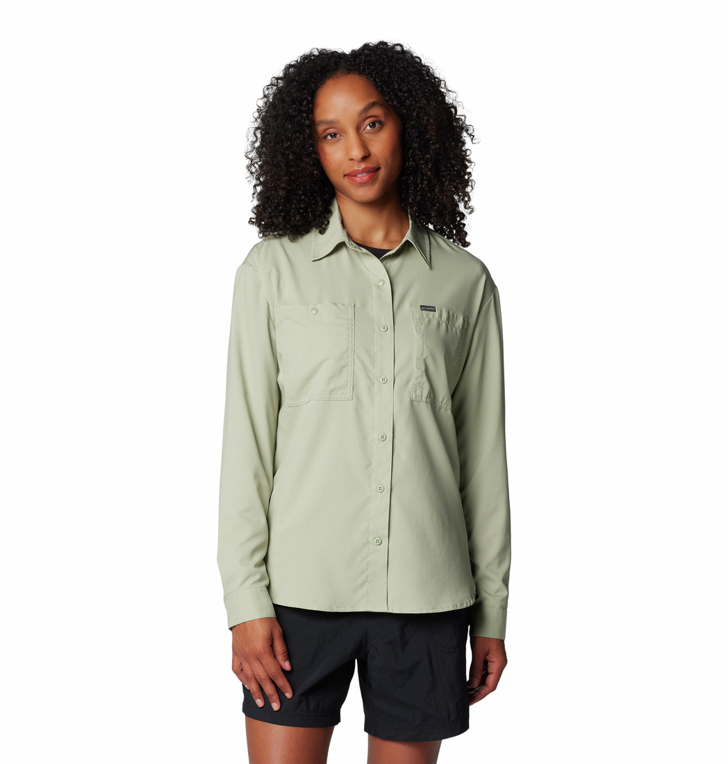 Une femme aux cheveux bouclés porte une chemise boutonnée à manches longues vert clair avec des poches sur la poitrine et un short noir, debout sur un fond blanc uni.
