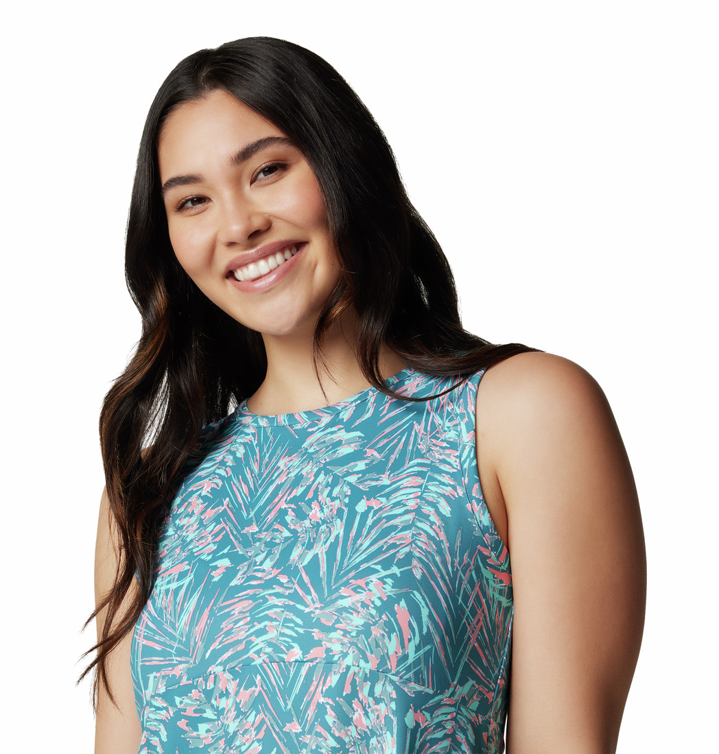 Une femme souriante aux longs cheveux noirs porte un haut sans manches à motifs bleus et roses. Elle se tient debout sur un fond blanc uni.