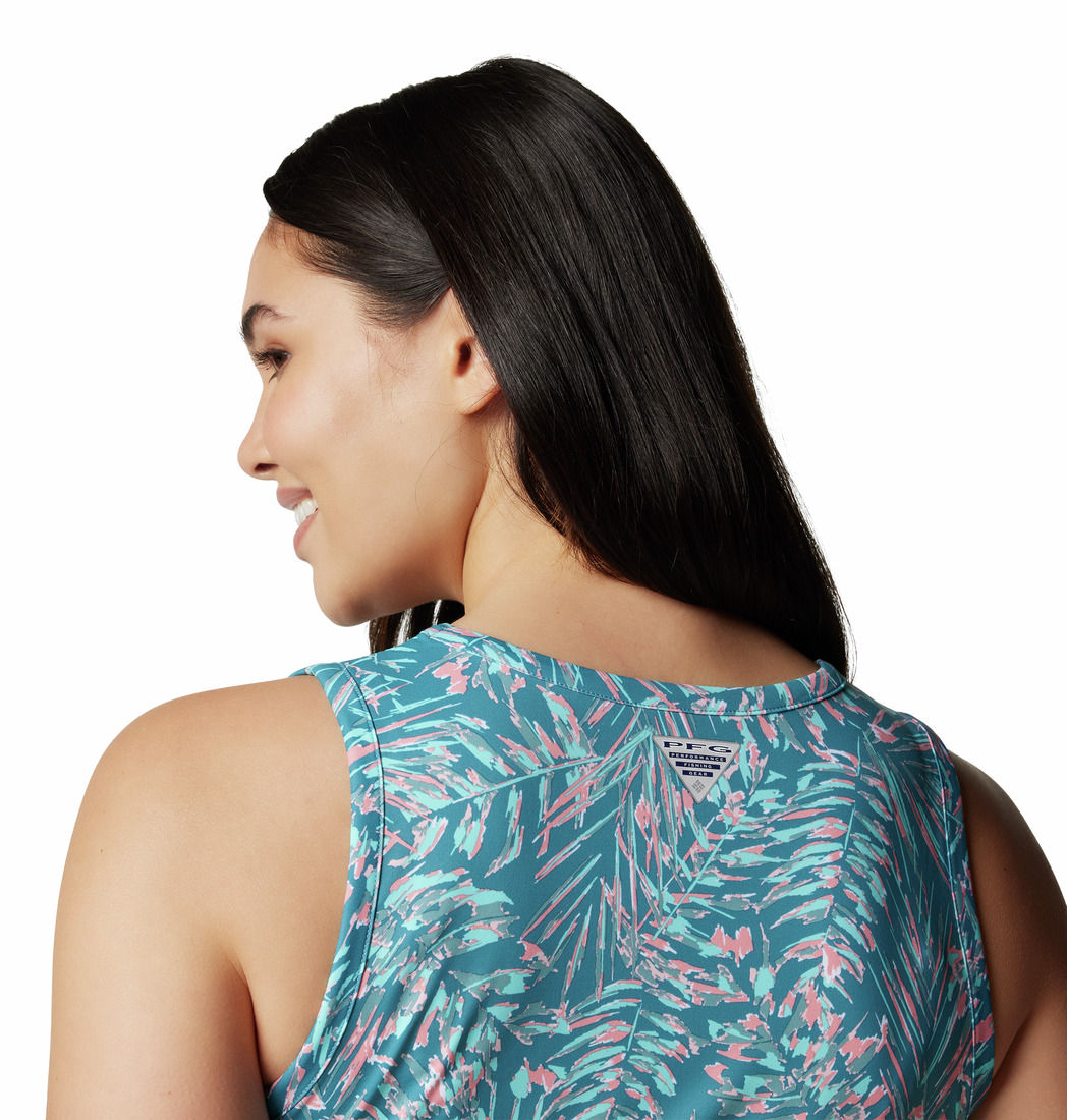 Une femme aux longs cheveux noirs sourit et regarde de côté, vêtue d'un haut sans manches bleu et rose à motifs tropicaux. La photo la montre de dos, épaules relevées, sur fond blanc.