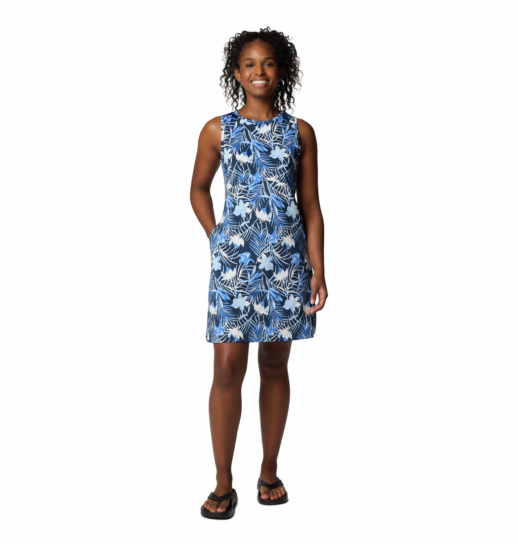 Une femme souriante porte une robe bleue sans manches, jusqu'aux genoux, à motif de feuilles tropicales blanches et bleu clair, et des sandales noires. Elle a les mains dans les poches et se tient debout sur un fond blanc.
