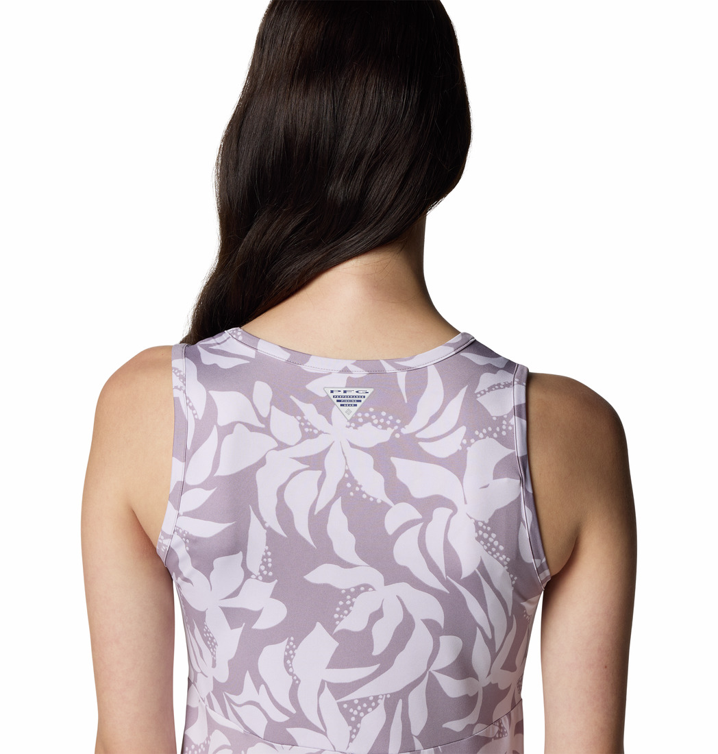 Une femme aux longs cheveux noirs porte un haut violet clair sans manches avec un motif floral abstrait blanc, photographié de dos sur un fond blanc.