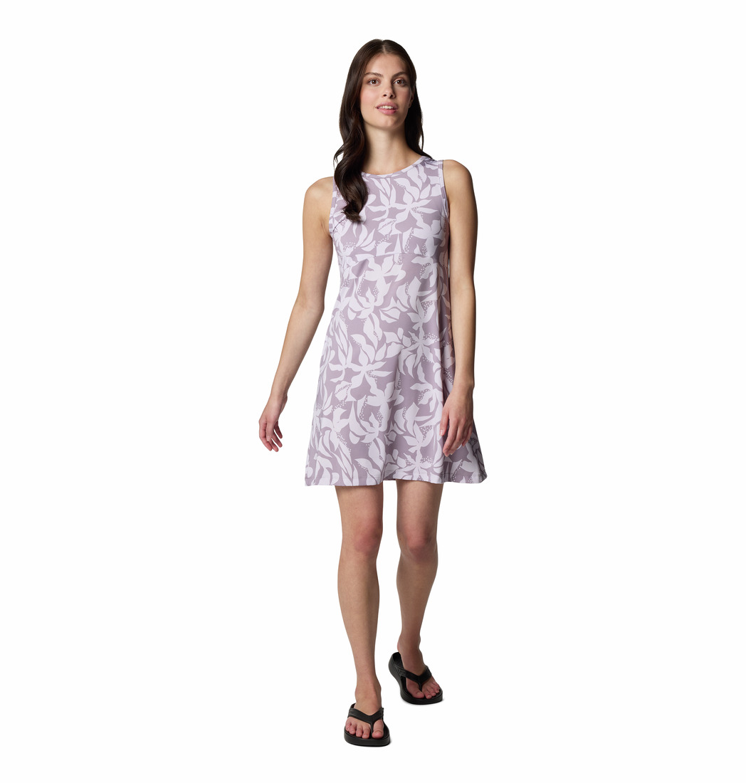 Une femme portant une robe sans manches de couleur lilas avec un motif de feuilles blanches, associée à des sandales noires, se tient debout sur un fond blanc uni.