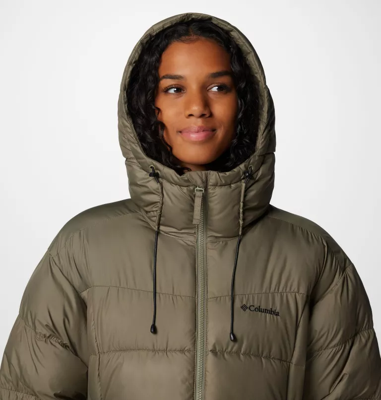 Une personne aux cheveux bouclés portant une doudoune Columbia vert olive à capuche sourit légèrement. Le fond est blanc uni.