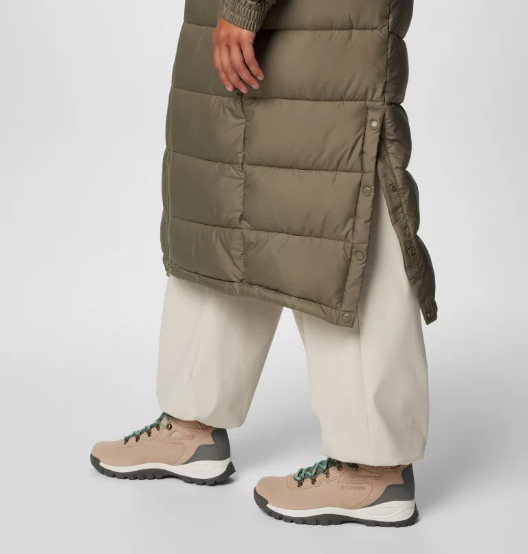Une personne portant un long manteau vert olive et un pantalon large ivoire se tient debout, les jambes croisées. Elle porte des bottes de randonnée beiges à lacets verts. L'arrière-plan est uni.