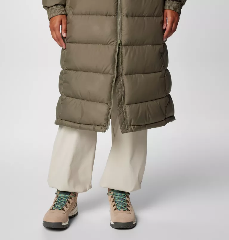 Une personne porte une longue doudoune vert olive, un pantalon large de couleur claire et des bottes de randonnée beiges à lacets bleu sarcelle sur un fond uni. L'accent est mis sur la moitié inférieure de sa tenue.