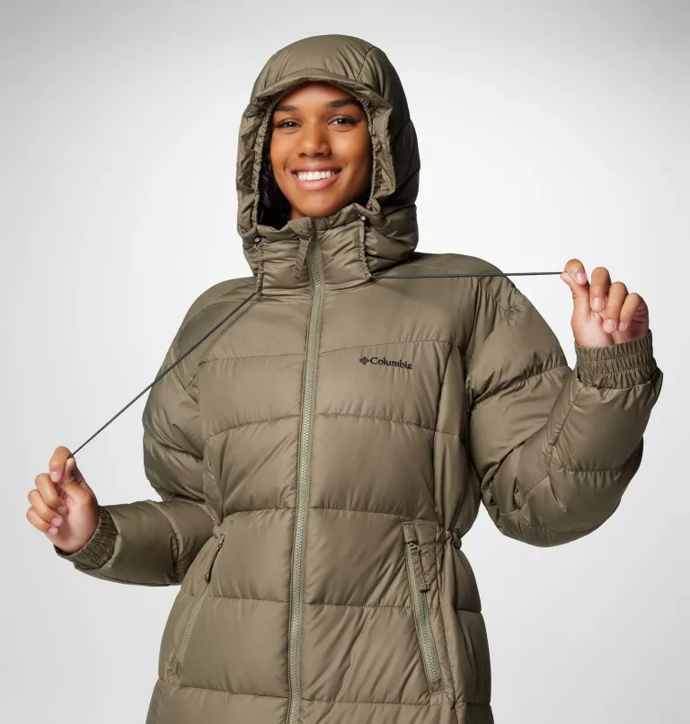 Une personne sourit tout en portant une veste Columbia vert olive à capuche bouffante, ajustant les cordons de la capuche. La veste comporte une fermeture éclair sur le devant et des poches latérales. L'arrière-plan est un simple dégradé de gris.