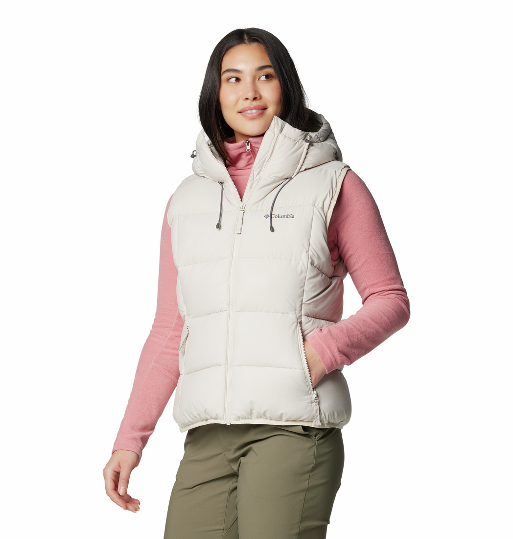 Une femme sourit en se tenant légèrement de côté. Elle porte la veste PINE LAKE II INSULATED VEST en beige clair avec une capuche et des cordons de serrage, superposée à un haut à manches longues rose et à un pantalon vert olive.