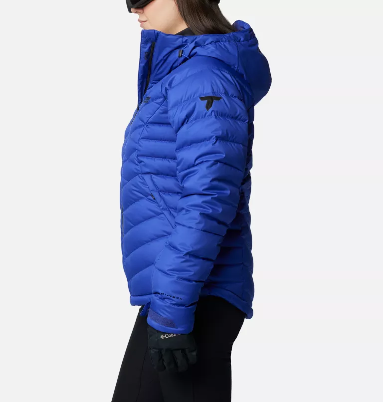 Vue latérale d'une personne portant une veste d'hiver bleu vif et bouffante avec une capuche, des gants noirs et un pantalon foncé. La veste a un logo discret sur la manche. Le fond est blanc uni.