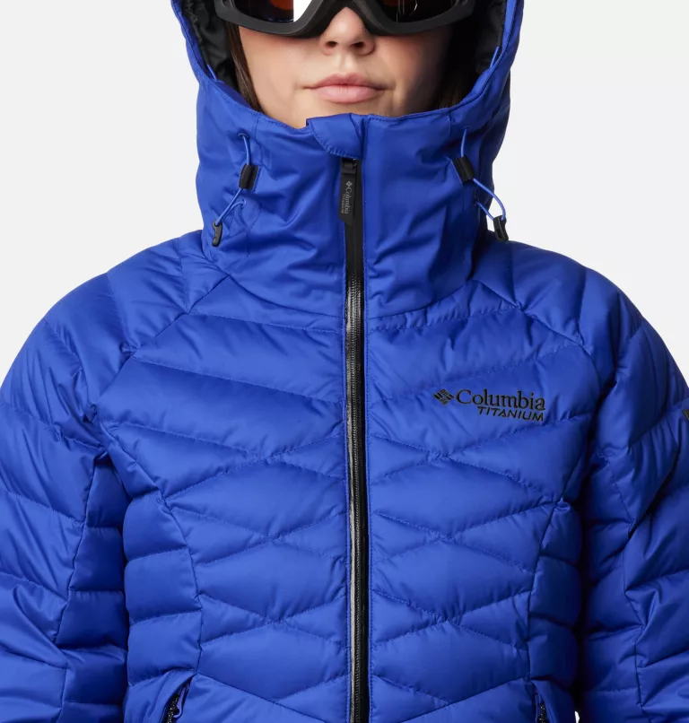 Une personne porte une doudoune Columbia Titanium bleu vif à capuche et des lunettes de ski. La veste a une fermeture éclair sur le devant et des coutures visibles créant un motif. Le fond est blanc.