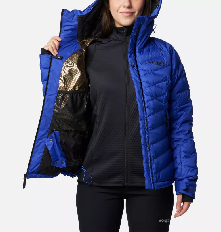 Une personne porte une veste isolante bleu vif avec des accents noirs, partiellement ouverte pour montrer une couche intérieure noire. La veste porte le logo Columbia sur la poitrine. La personne tient la veste ouverte d'une main.