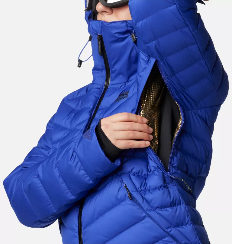 Une personne portant une veste d'hiver isolante bleue ajuste la fermeture éclair de ventilation latérale. La veste a un col haut et une capuche avec cordons de serrage. Seul le haut du corps est visible sur un fond blanc uni.