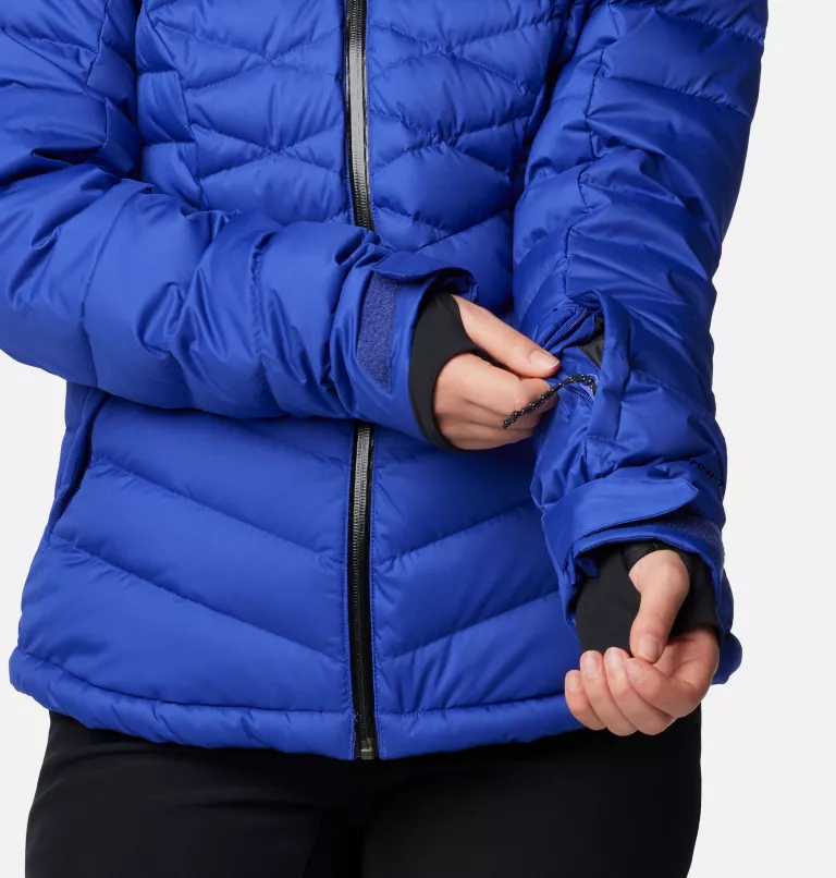 Une personne portant une doudoune bleu vif ajuste une poche zippée sur la manche. La veste présente des motifs matelassés et la personne est vêtue d'une couche de base noire en dessous, l'accent étant mis sur les détails des manches.