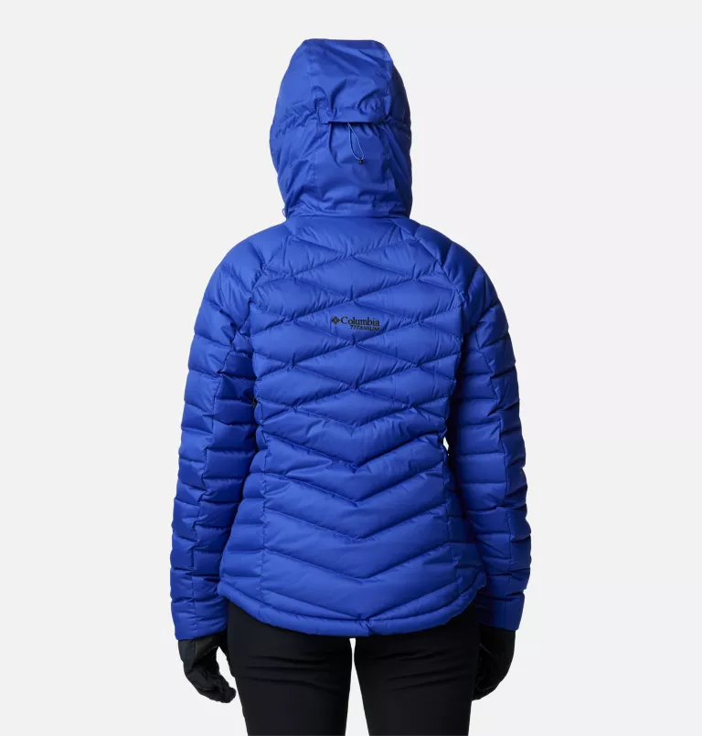 L'image montre une personne portant une doudoune bleu vif avec une capuche. La veste a un motif matelassé en zigzag. La personne est tournée vers l'extérieur, debout sur un fond gris clair uni, et porte un pantalon et des gants noirs.