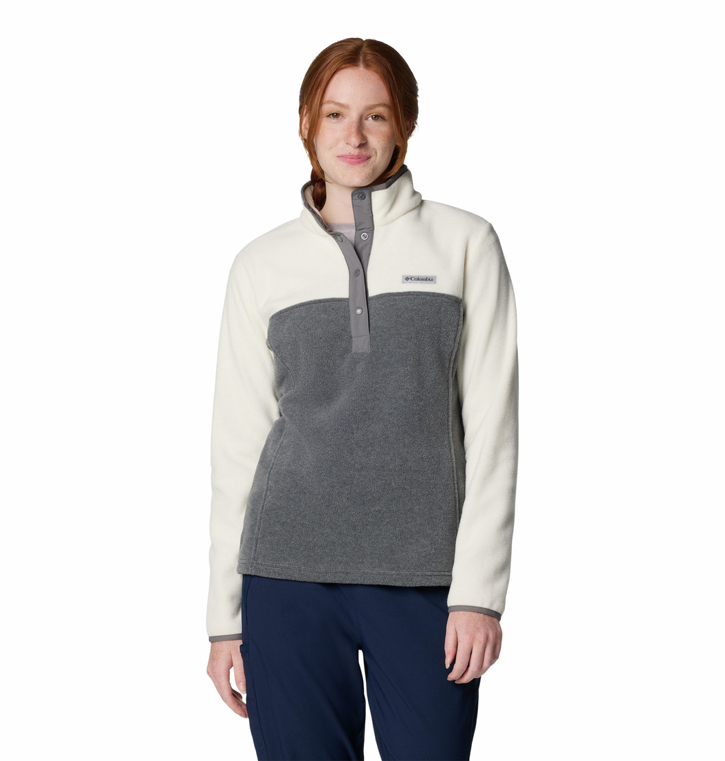 Une femme aux cheveux roux, vêtue d'un pull polaire gris clair et foncé et d'un pantalon bleu marine, se tient debout sur un fond blanc uni, souriant doucement à la caméra.