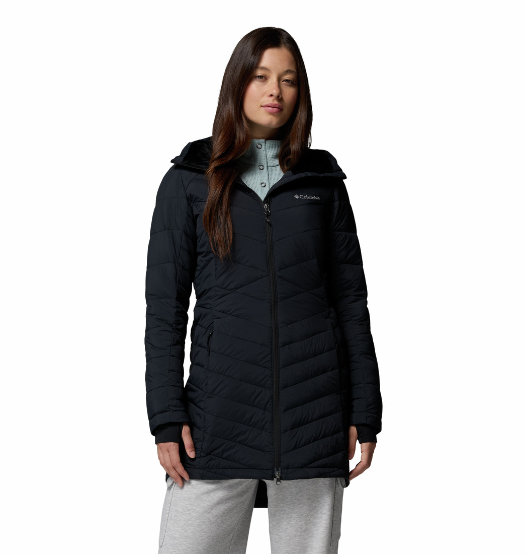 Une femme portant une longue veste noire à capuche, un haut bleu clair et un pantalon gris clair se tient debout, de face, avec une expression neutre, sur un fond blanc uni.