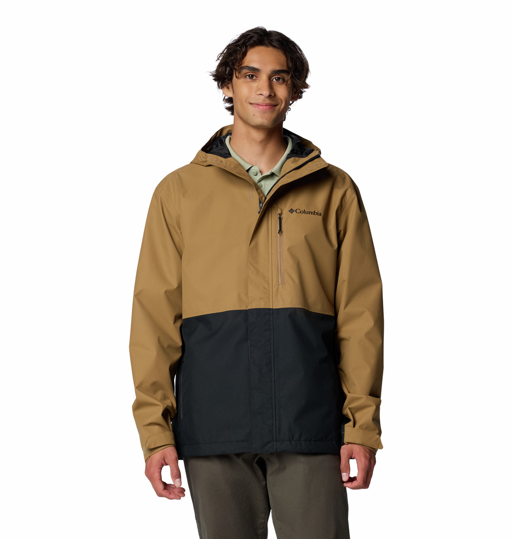 Un jeune homme aux cheveux bruns ondulés porte la veste HIKEBOUD II JACKET, un modèle à capuche bicolore avec le haut brun et le bas noir, une poche de poitrine zippée et le logo "Columbia" sur le côté gauche. Il est tourné vers l'avant et sourit légèrement.
