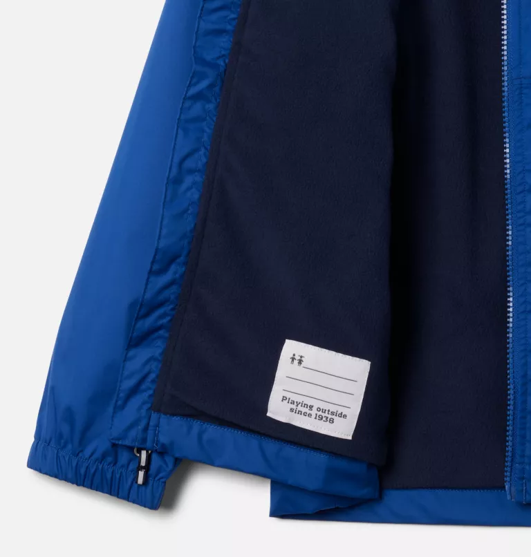 Gros plan sur une veste bleue partiellement ouverte, révélant une doublure sombre. Une étiquette blanche à l'intérieur indique « Jouer dehors depuis 1938 ». La manche est dotée d'un poignet élastique. L'image met en valeur les détails intérieurs de la veste.