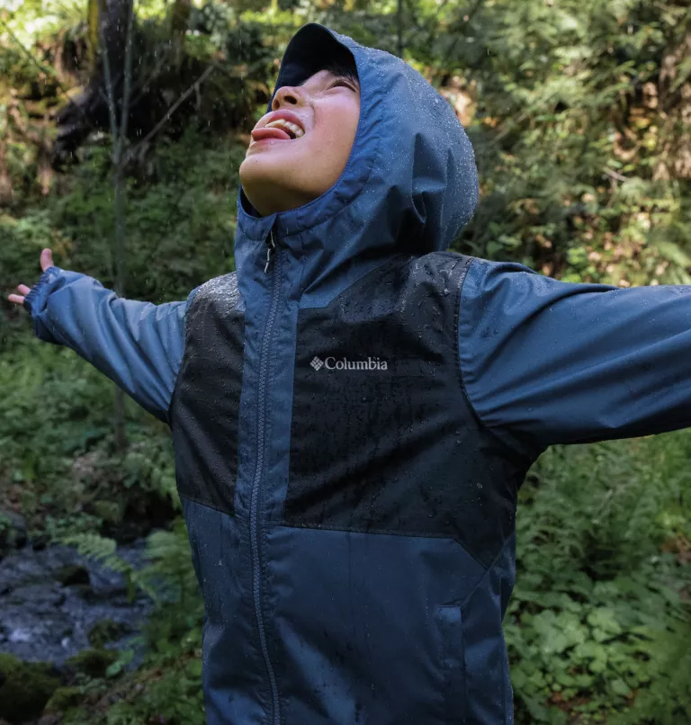 Un homme vêtu d'un imperméable bleu se tient debout, les bras tendus et la tête en arrière, attrapant les gouttes de pluie avec sa langue. Il est entouré d'un feuillage vert luxuriant dans un décor forestier.