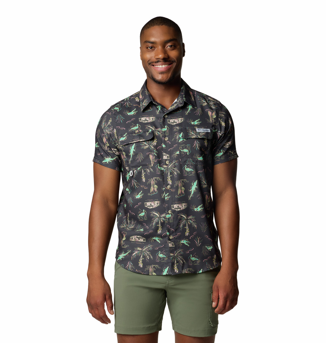 Un homme sourit, vêtu d'une chemise à manches courtes boutonnée à imprimé tropical et d'un short vert. Il se tient debout, le visage tourné vers l'avant, sur un fond blanc uni.