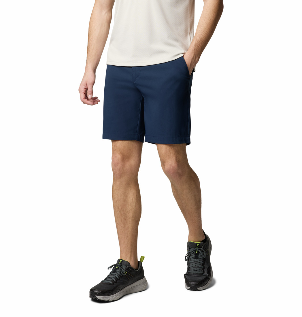 Une personne portant une chemise blanche à manches courtes, un short bleu marine et des chaussures de sport noires se tient debout avec une main dans sa poche sur un fond blanc uni.