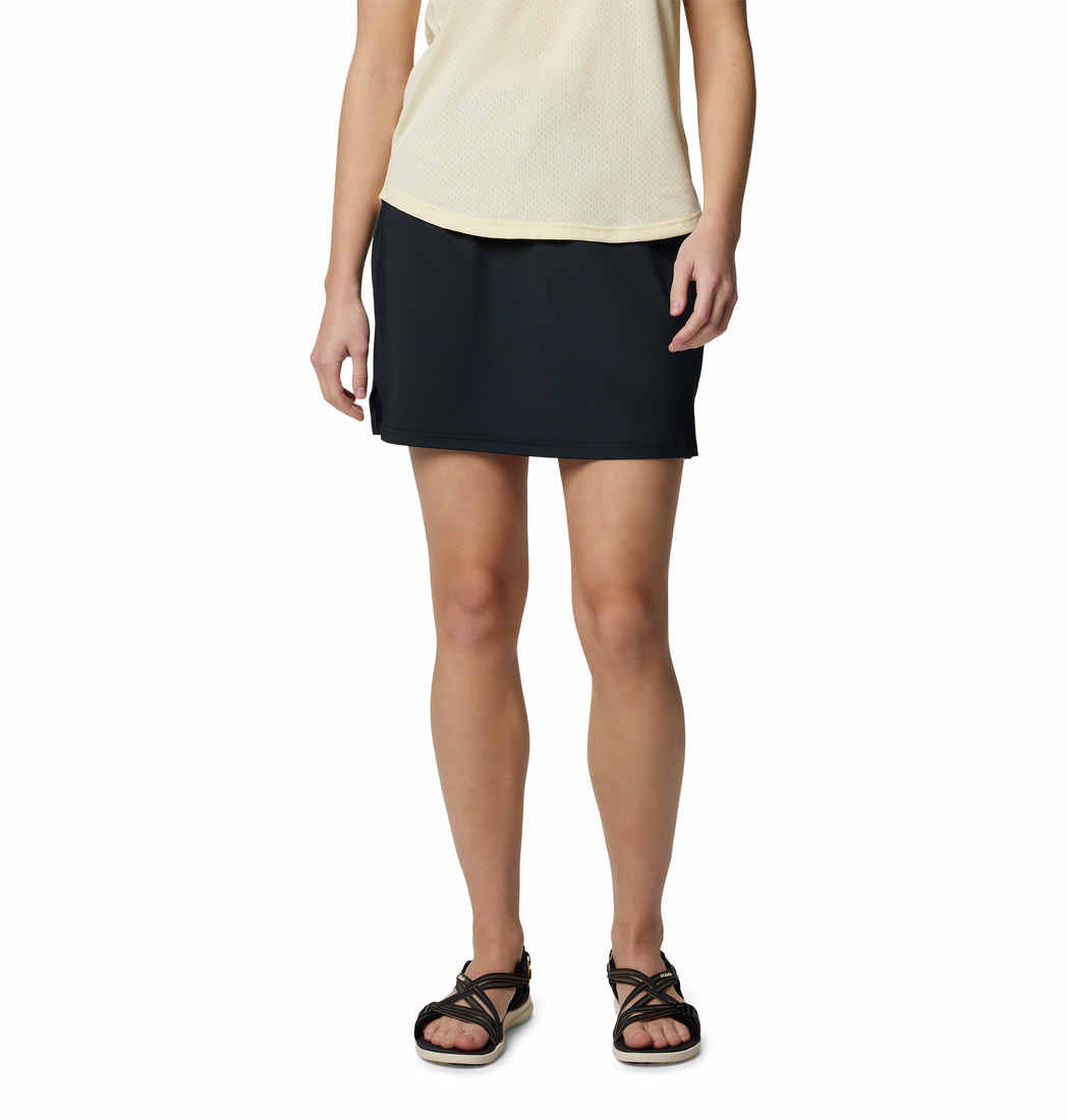 Une personne portant un haut beige sans manches, une jupe noire et des sandales noires se tient debout sur un fond blanc. L'image est recadrée au niveau des épaules, ne laissant apparaître que le bas du torse et les jambes.
