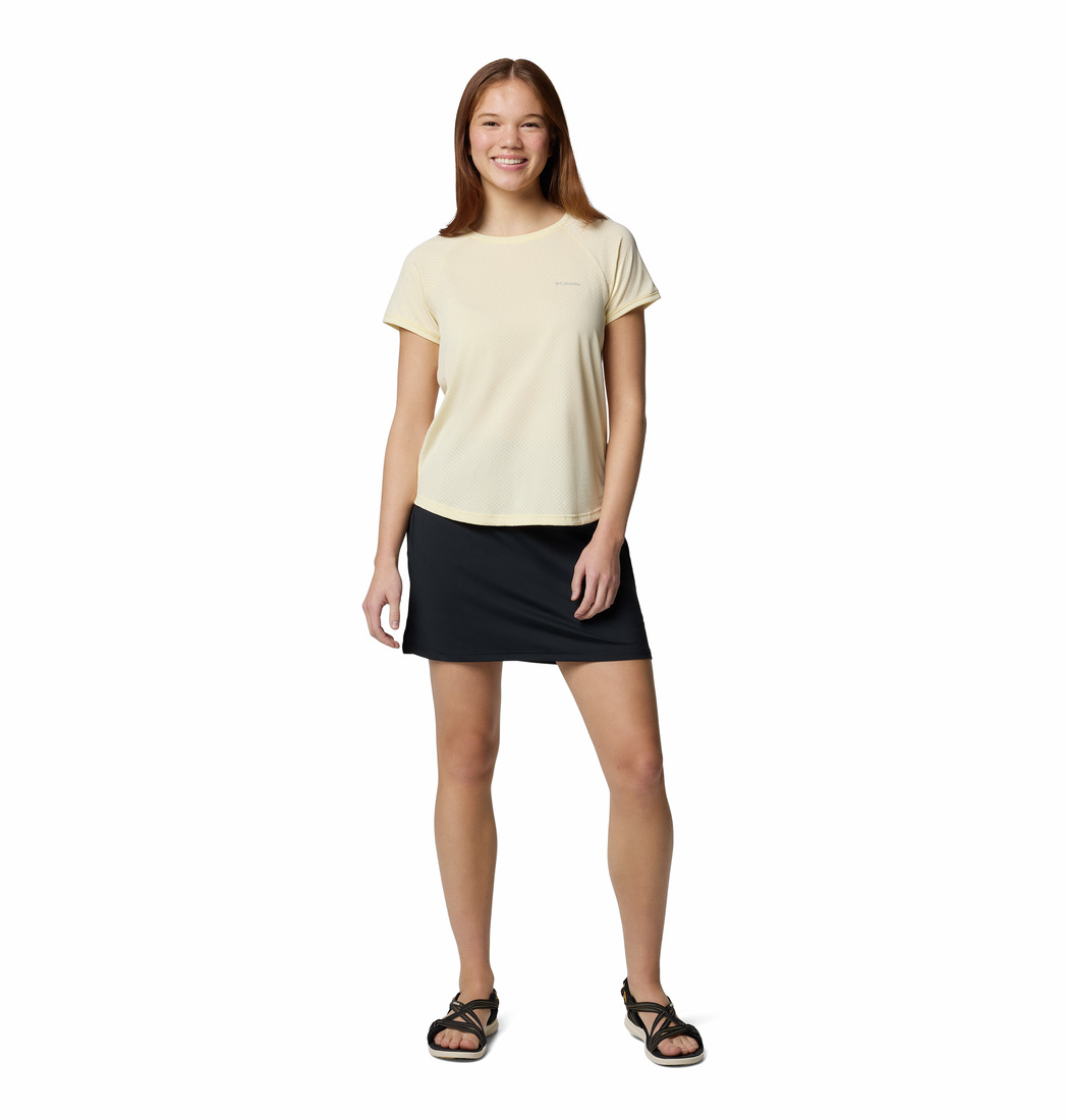 Une femme souriante se tient debout sur un fond blanc, vêtue d'un t-shirt jaune clair à manches courtes, d'une jupe noire et de sandales noires. Elle a les cheveux bruns et raides et une main posée sur le côté.