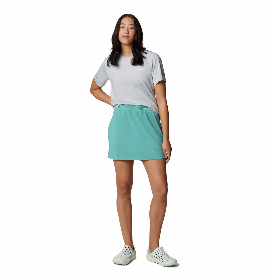Une femme se tient debout sur un fond blanc, vêtue d'un t-shirt gris clair, d'une mini-jupe bleu canard et de mocassins blancs. Elle a de longs cheveux noirs et une pose décontractée.