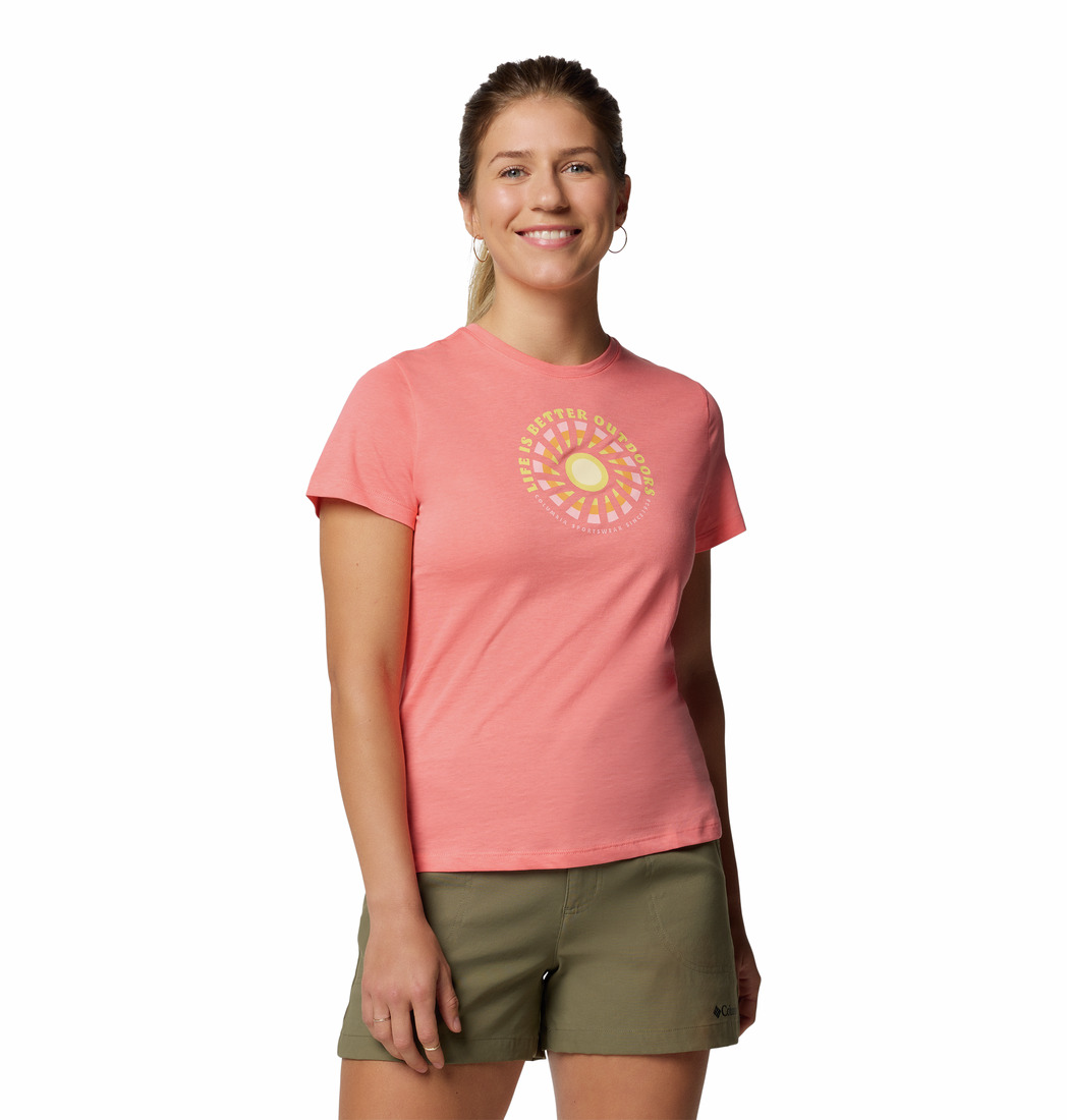 Une femme souriante porte un t-shirt rose clair orné d'un motif circulaire jaune et blanc sur le devant, ainsi qu'un short vert olive. Le fond est blanc uni.