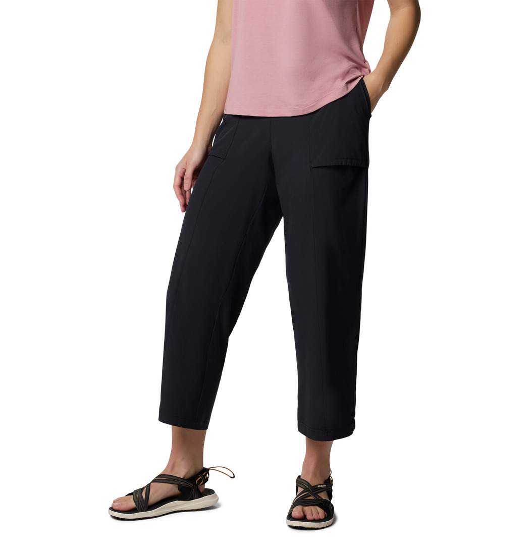 Une personne portant un haut rose à manches courtes, un pantalon noir court à poches et des sandales noires à semelles blanches, debout, une main dans une poche, sur fond blanc. Sa tête n'est pas visible sur l'image.