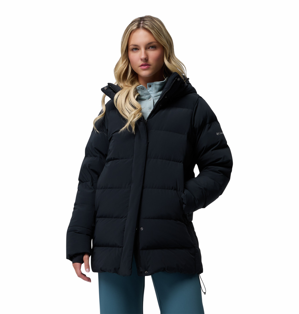 Une femme aux longs cheveux blonds porte un blouson noir par-dessus un chemisier bleu clair et un pantalon bleu, debout, les mains dans les poches, sur un fond blanc uni.